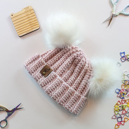 A light pink ribbed knit beanie with two large white faux-fur pom-poms and a small suede logo patch, arranged on a white background with craft props including scissors and stitch markers.