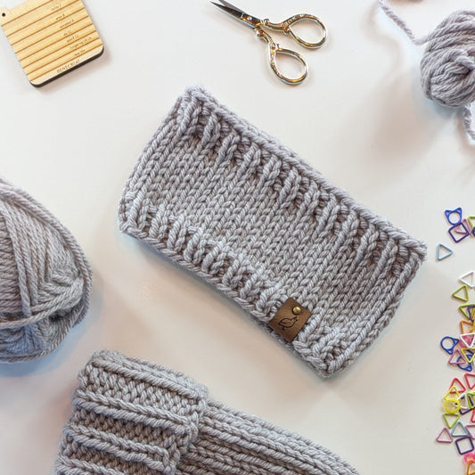 A light gray knitted headband with a textured rib-stitch pattern and a small brown leather tag, displayed on a white surface with knitting supplies including yarn balls, scissors, and colorful stitch markers.