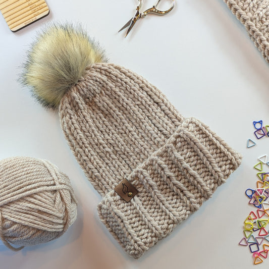 A chunky knit beanie hat in a warm oatmeal beige color, featuring a large fluffy pom-pom and a ribbed cuff with a small suede logo tag. The hat is styled in a flat-lay arrangement with a ball of matching yarn, gold scissors, and colorful stitch markers on a white background.