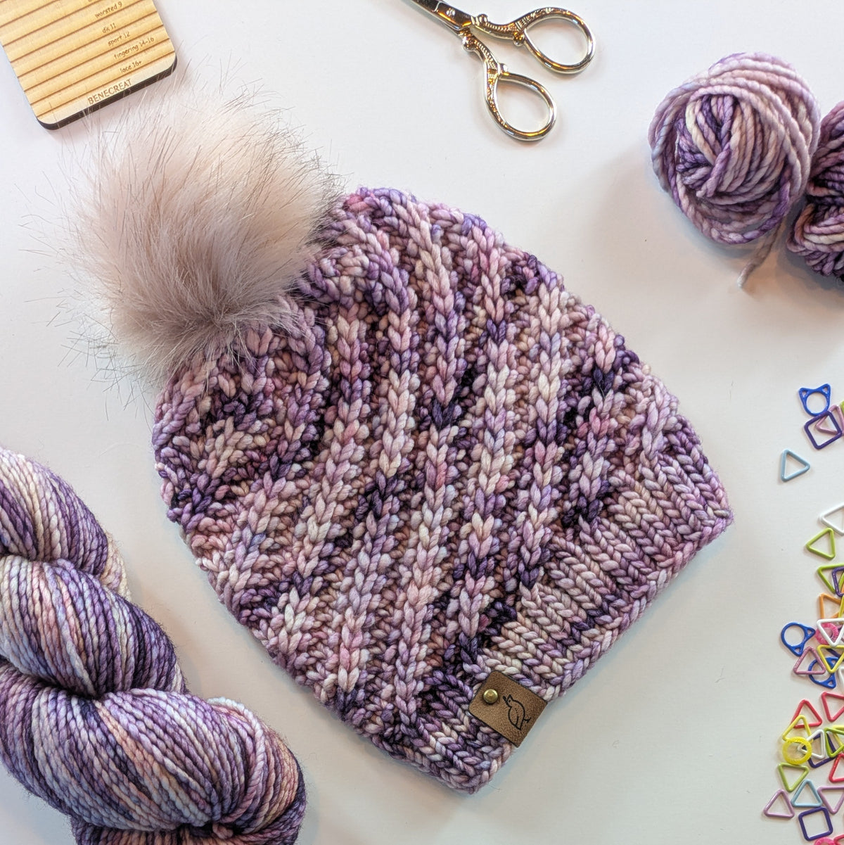 A hand-knit beanie hat with a fluffy pom-pom, laid flat on a white surface. The hat features a variegated pattern of dusty purple, mauve, and pink yarn.