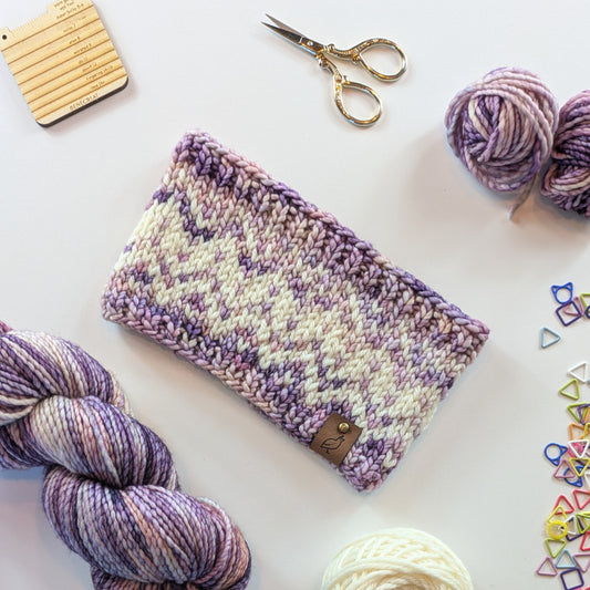 A hand knit fair isle ear warmer headband in a variegated pattern of cream with purple and pink yarn, styled in a flat-lay with knitting supplies on a white background.