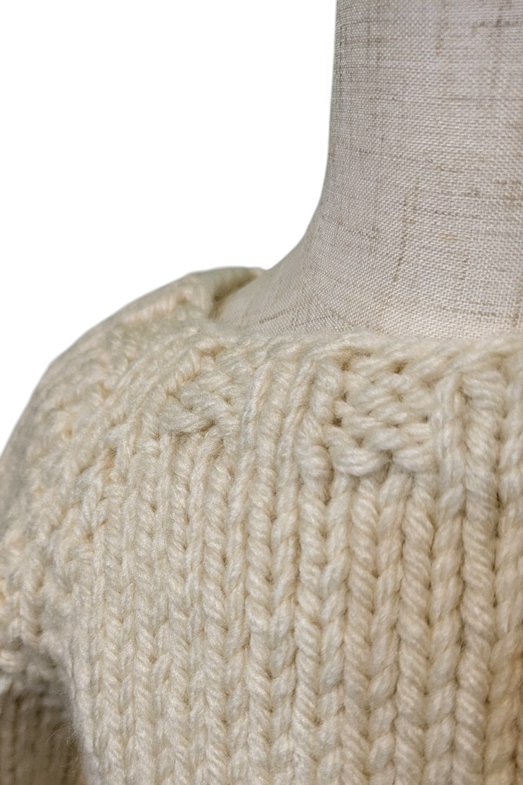 Close-up of a chunky knit sweater in a warm cream or off-white color, displayed on a linen mannequin torso against a white background.
