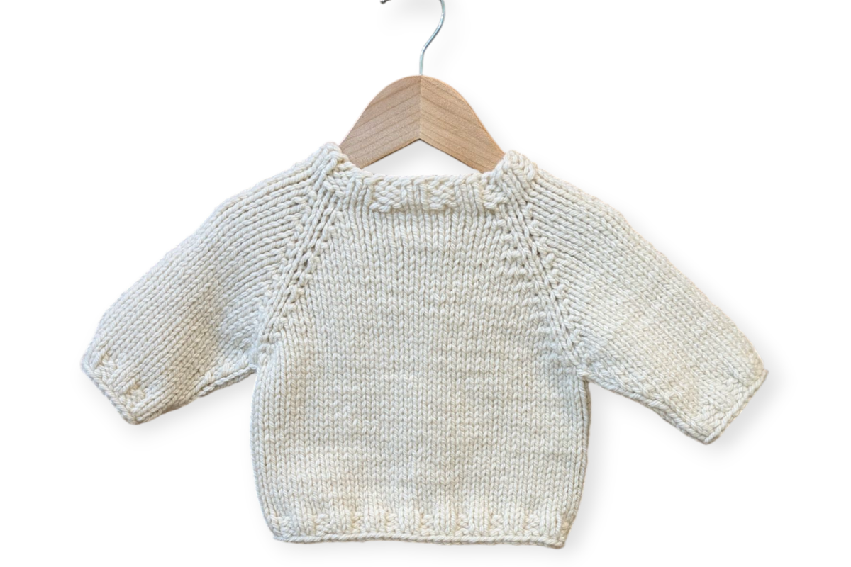 A cream-colored hand-knit baby sweater with long sleeves and crew neck, hanging on a wooden hanger against a plain white background.