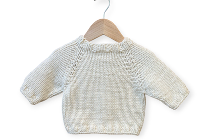 A cream-colored hand-knit baby sweater with long sleeves and crew neck, hanging on a wooden hanger against a plain white background.