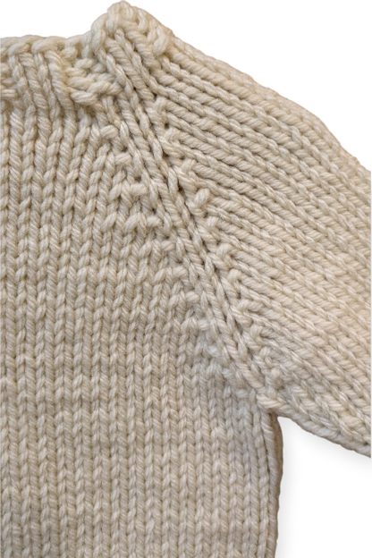 Close-up of a cream-colored, chunky hand-knit sweater showing the detailed ribbing and sleeve construction against a plain white background.