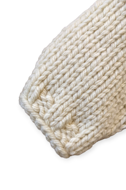 A close-up of a chunky hand-knit sweater sleeve, in a warm cream or off-white color, showing the ribbed cuff detail against a plain white background.