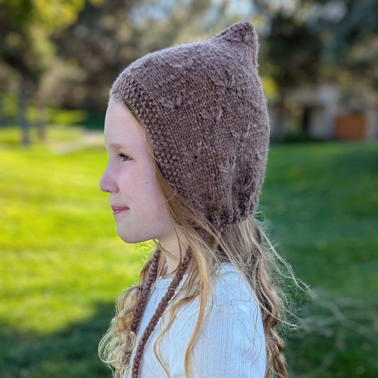 A young girl in profile wearing a brown bonnet with subtle dandelion pattern and drawstring, set against a blurred green grassy outdoor background.