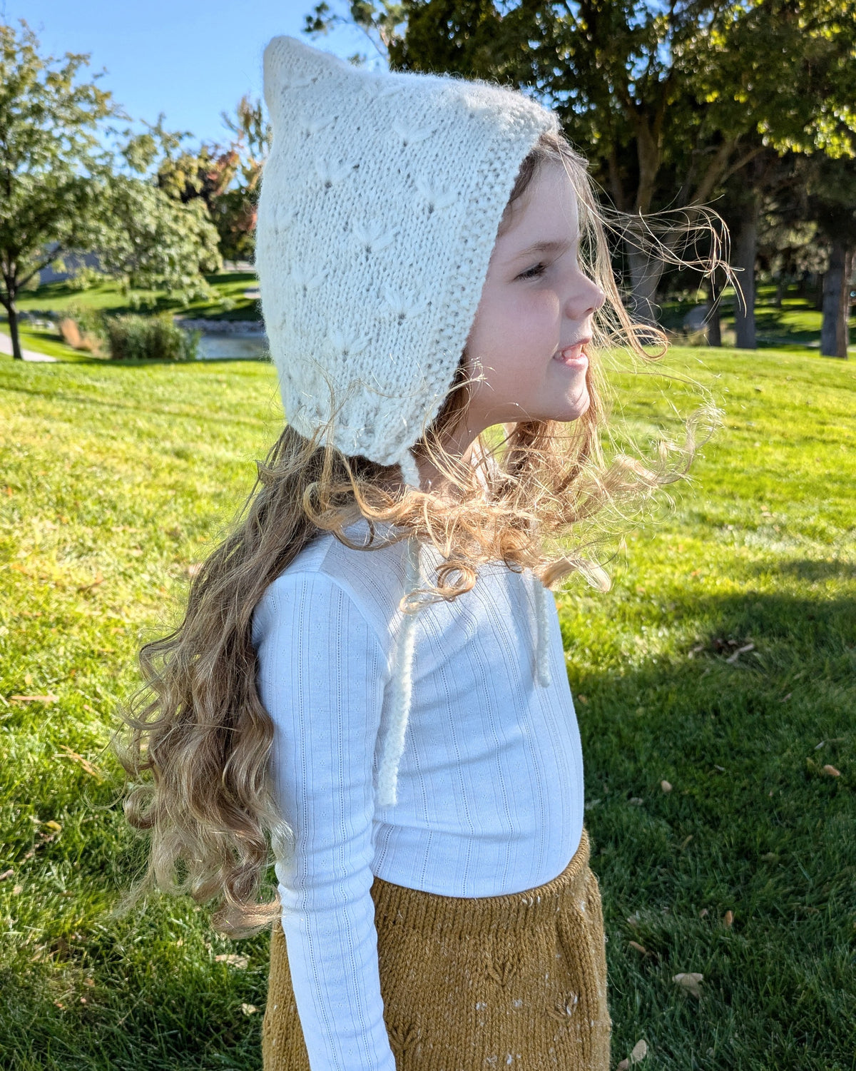 A young girl with long curly blonde hair is shown in profile outdoors on a grassy lawn, wearing a cream hand knit bonnet with subtle dandelion pattern and drawstring, a white pointelle long-sleeve top, and a gold hand knit skirt.