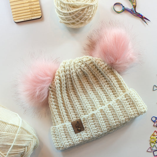 A cream-colored ribbed knit beanie hat with two fluffy light pink pom-poms, displayed on a light background with knitting supplies like yarn, scissors, and a wooden gauge tool.