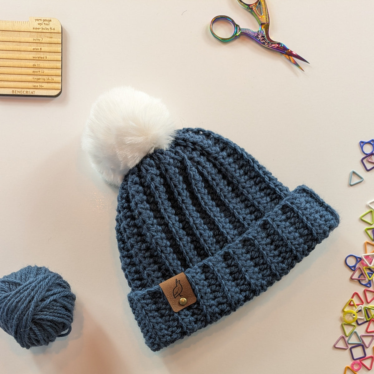 A blue ribbed knit beanie with a white pom-pom on top and a small brown leather logo tab on the cuff, arranged on a light background with yarn, scissors, a ruler, and colorful triangle stitch markers around it.