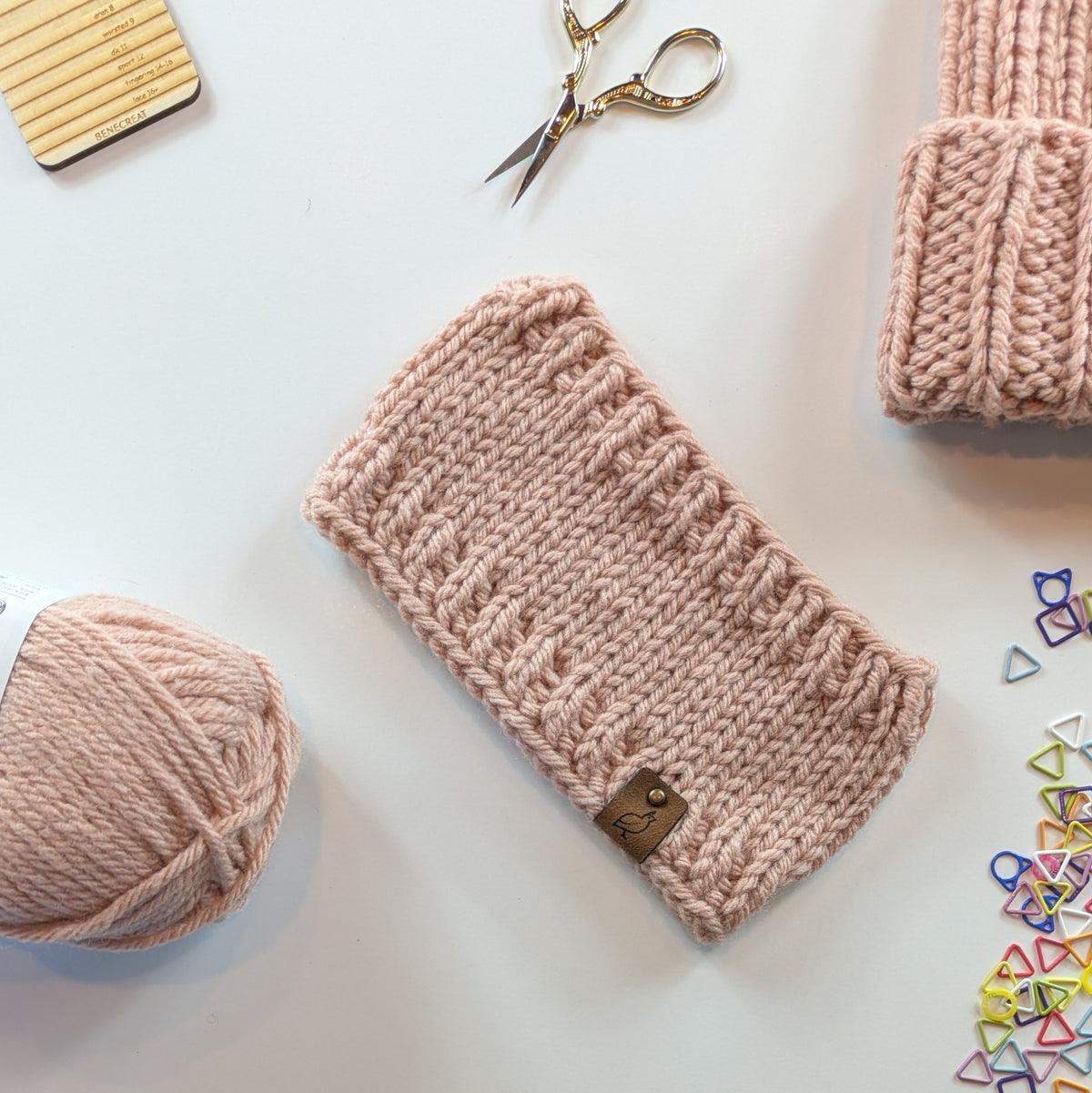 A pale blush pink knitted headband laid flat on a white surface, surrounded by knitting accessories including a ball of matching yarn, gold scissors, a folded matching knit hat, and colorful stitch markers.