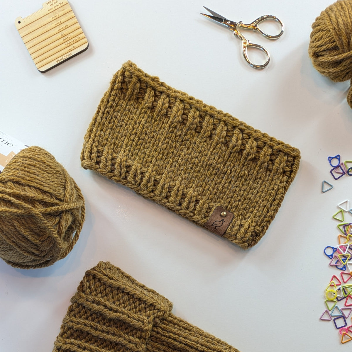 A flat-lay view of a hand-knit brown headband with a small leather brand tag, surrounded by knitting supplies including yarn balls, scissors, and colored stitch markers on a white background.