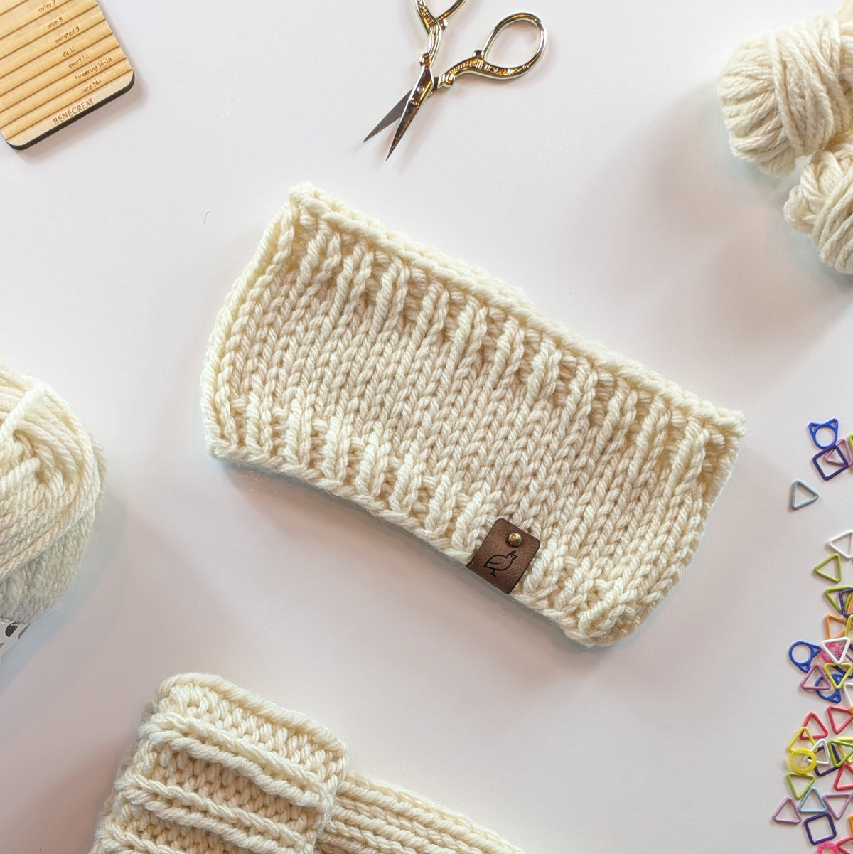 A cream-colored chunky knit headband with a small brown leather brand tag, displayed on a white background with knitting props like yarn, scissors, and stitch markers.