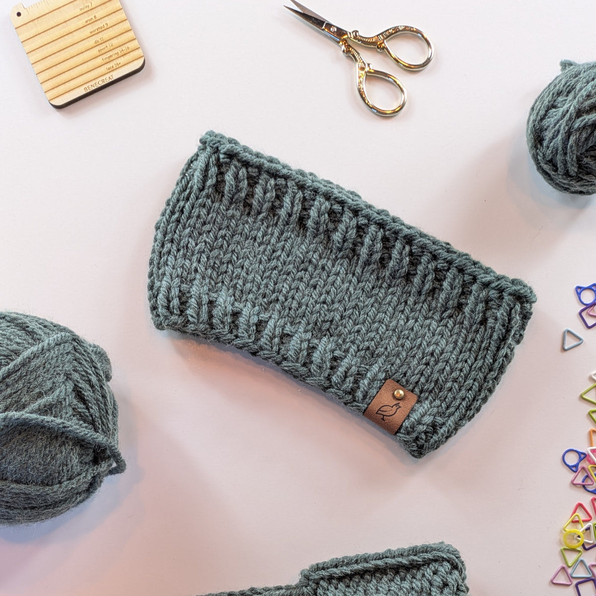A forest-green knitted headband with a small brown leather brand tab, styled in a flat-lay with yarn balls, gold scissors, and knitting tools on a white background.