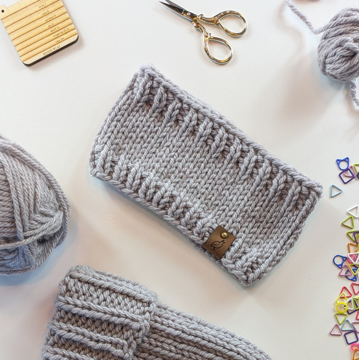 A light gray knitted headband with a textured rib-stitch pattern and a small brown leather tag, displayed on a white surface with knitting supplies including yarn balls, scissors, and colorful stitch markers.
