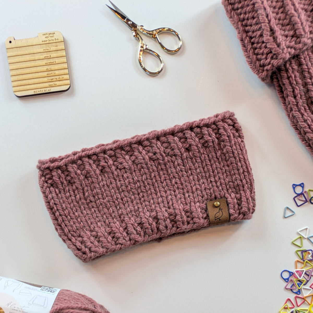 A dusty rose mauve chunky knit headband with a small brown leather brand tag, displayed in a styled flat-lay with knitting scissors, a wooden stitch-count card, and other knitting accessories on a white background.