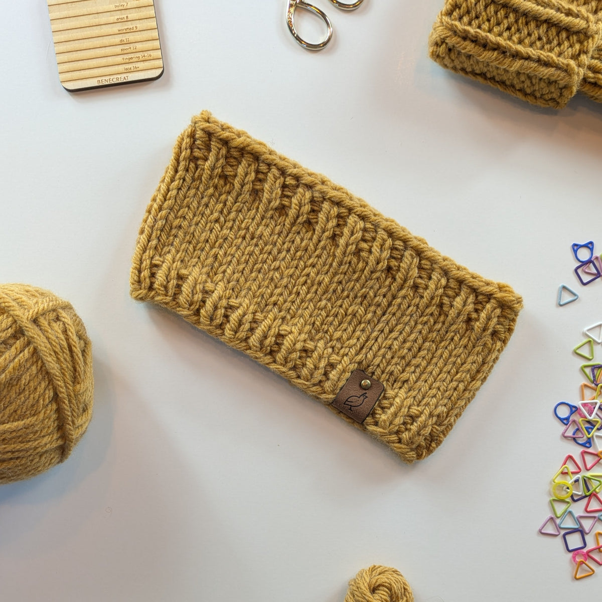 A mustard yellow knitted headband with a small brown leather logo tab, displayed in a flat-lay arrangement with knitting supplies on a white background.