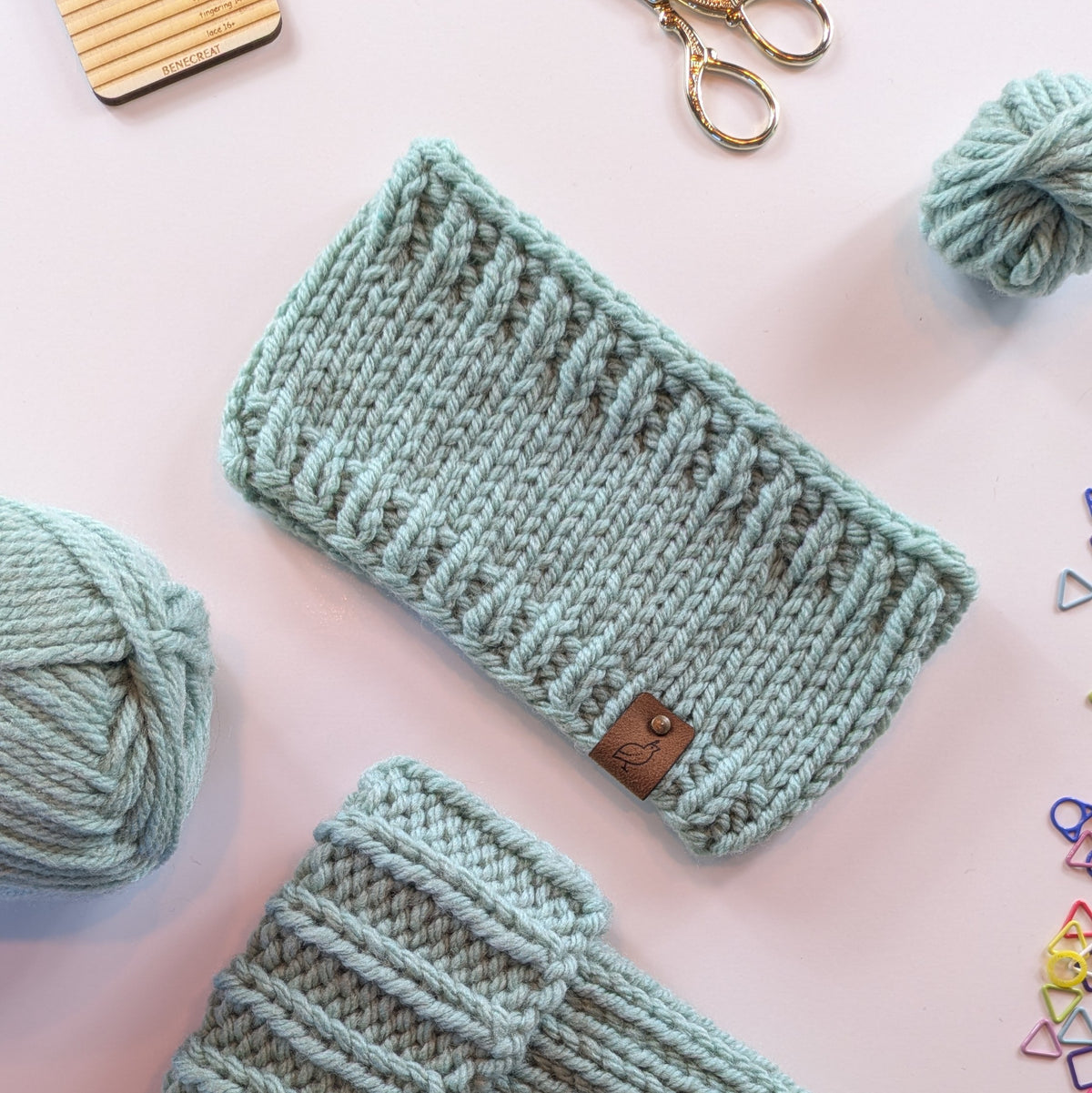 A mint green chunky knit beanie with a small brown leather brand tab, displayed on a light background with knitting items like yarn balls, scissors, and stitch markers.