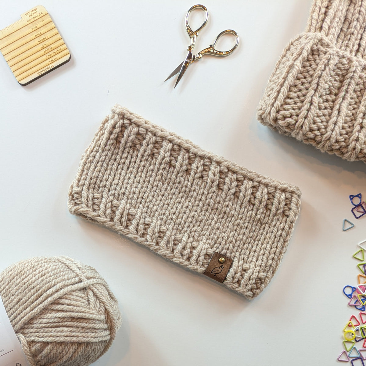 A beige chunky-knit fabric headband with a small brown leather brand tab, styled in a flat lay with a ball of matching yarn, gold scissors, and other knitting accessories on a white background.