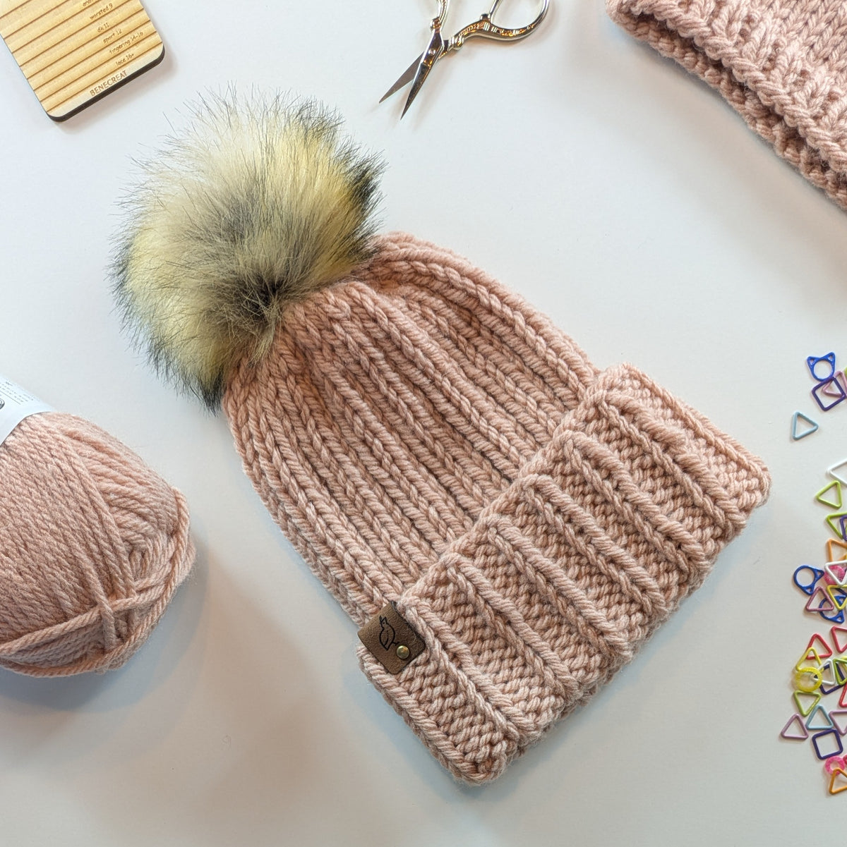 A chunky knit beanie in a dusty pink color with a large fluffy faux-fur pom-pom in beige, brown, and black tones. The beanie is styled in a flat-lay arrangement on a white surface alongside a ball of matching pink yarn, scissors, a wooden gauge card, and colorful stitch markers.