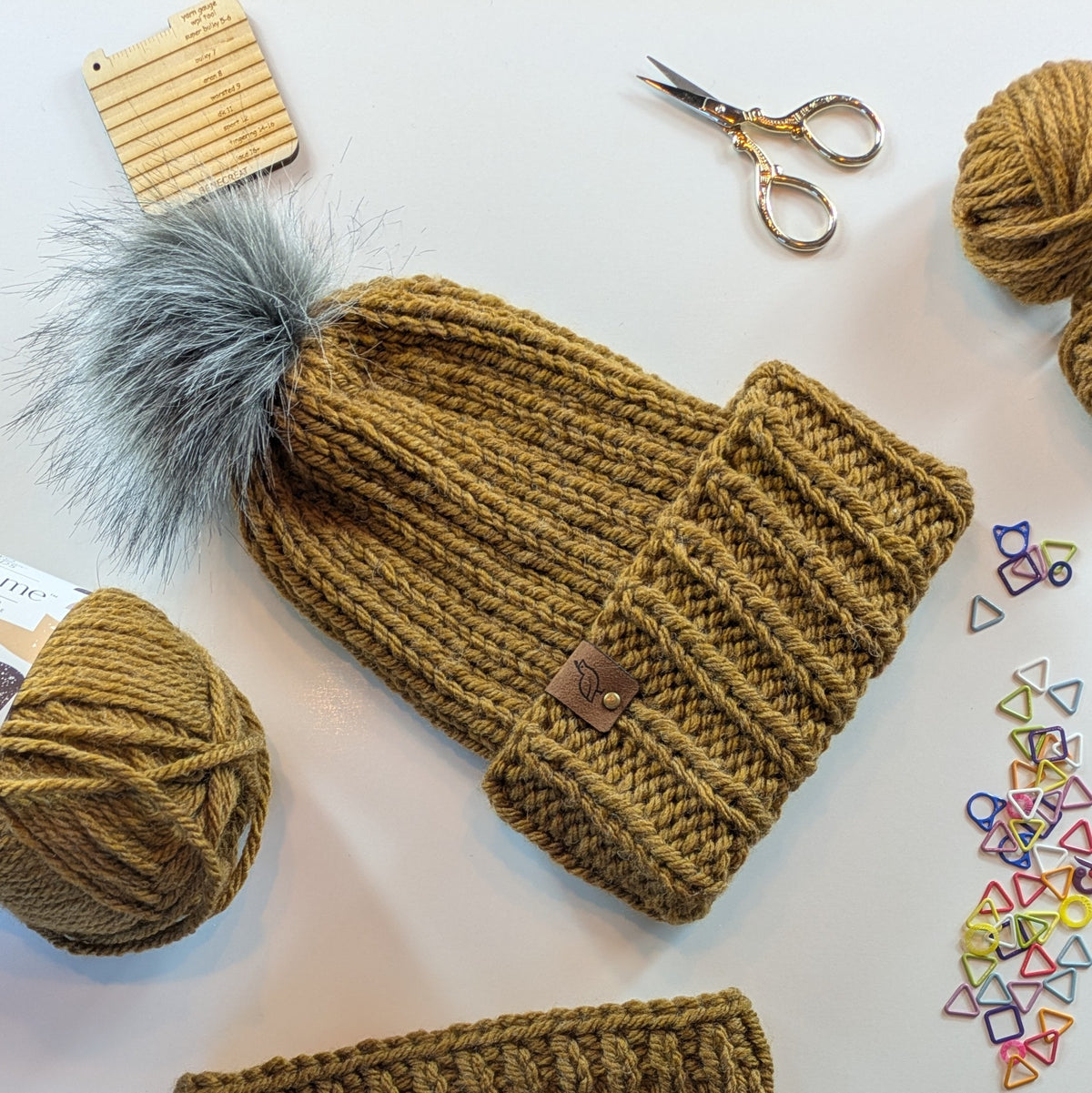A chunky knit beanie in a warm camel brown color, styled in a flat-lay with a gray faux-fur pom-pom, yarn balls, scissors, and small colorful metal rings on a white background.