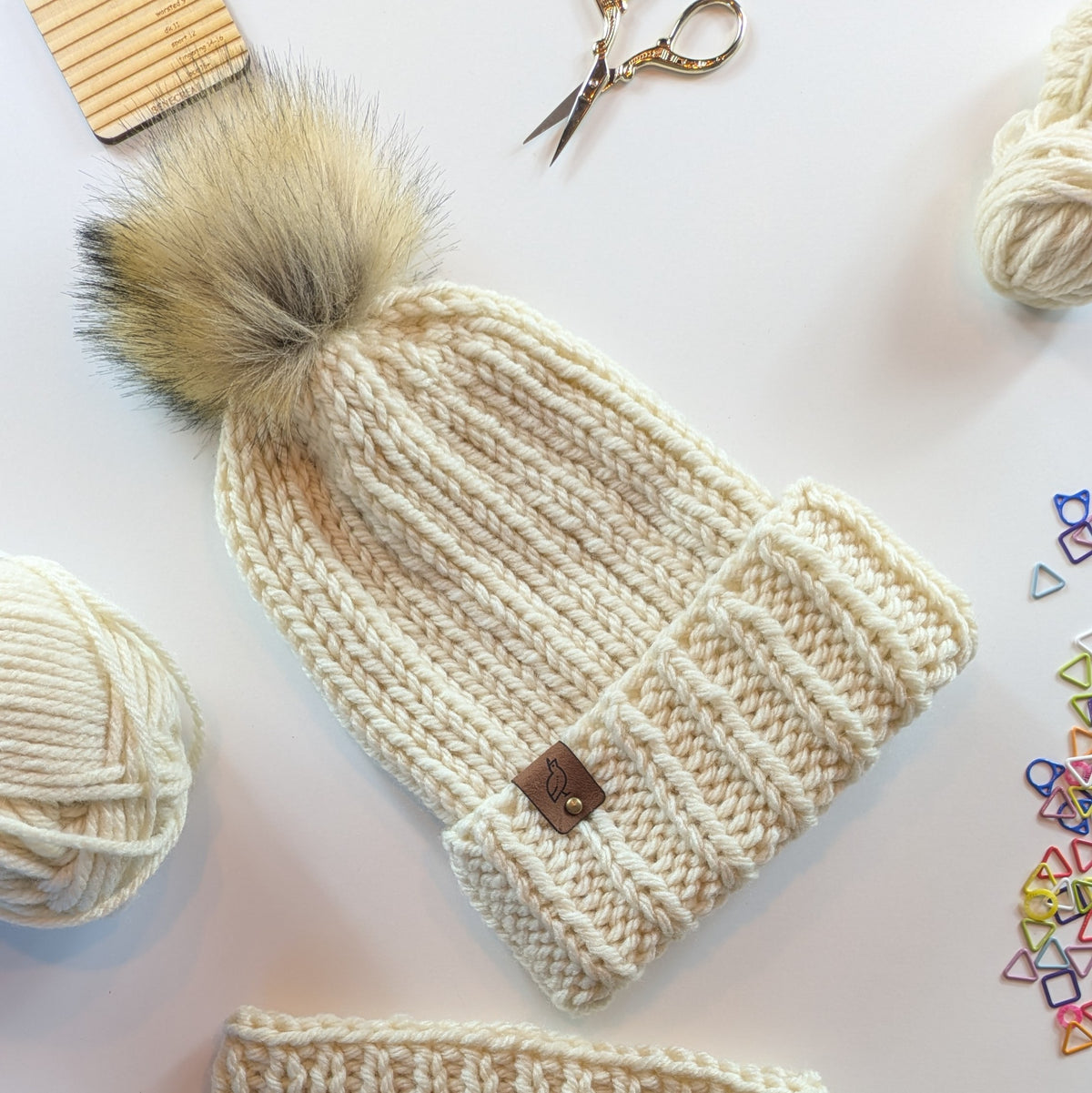 Cream-colored chunky knit beanie hat with a large fluffy pom-pom and a small brown leather brand tag, styled in a flat lay with yarn balls, scissors, and colorful stitch markers on a white background.