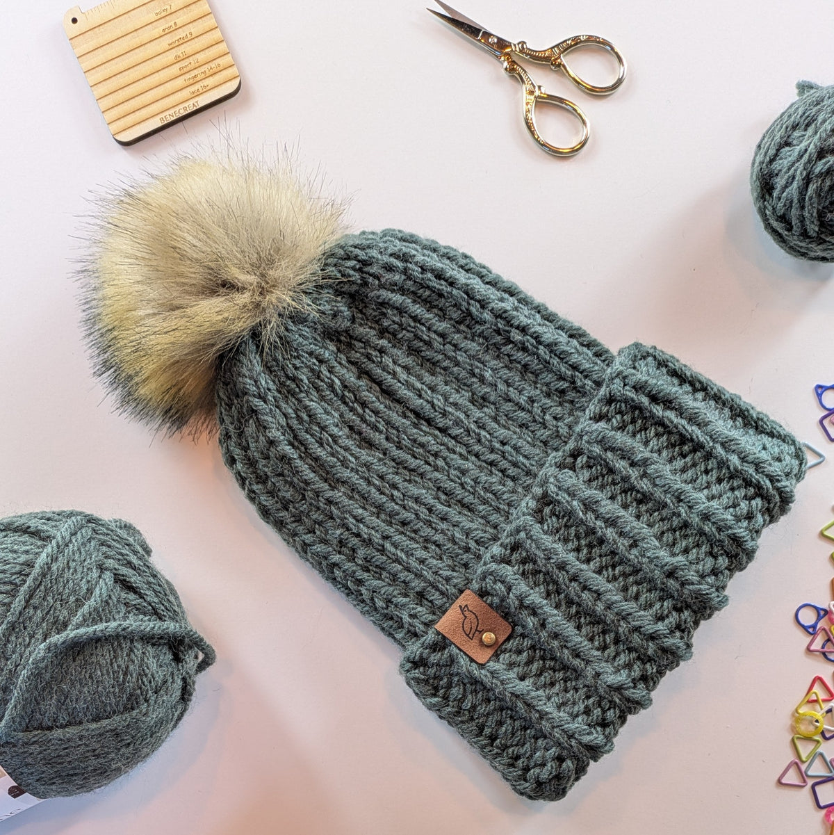 A chunky knit beanie in muted forest green with a large fluffy pom-pom, laid flat on a light background with yarn balls, scissors, and small colorful accessories around it.