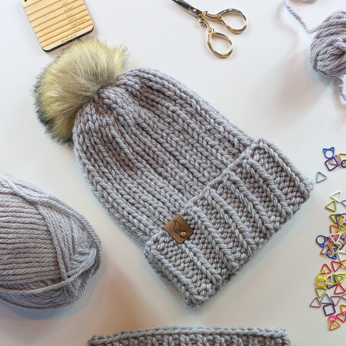 A light gray chunky knit beanie with a fluffy pom-pom in beige and gray, styled in a flat lay with yarn balls, scissors, colorful triangle stitch markers, and a wooden gauge tool on a white background.