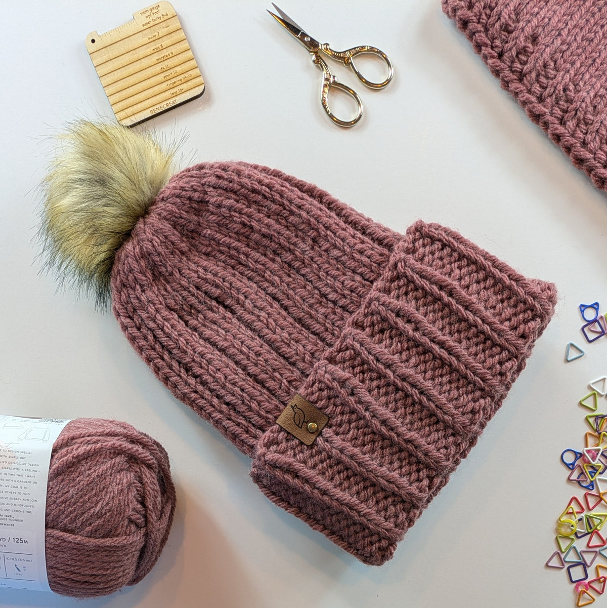 A chunky knit pom-pom beanie in dusty rose mauve, styled in a flat lay with a matching ball of yarn, gold scissors, a wooden measuring tool, and small colorful geometric stitch markers on a white background.