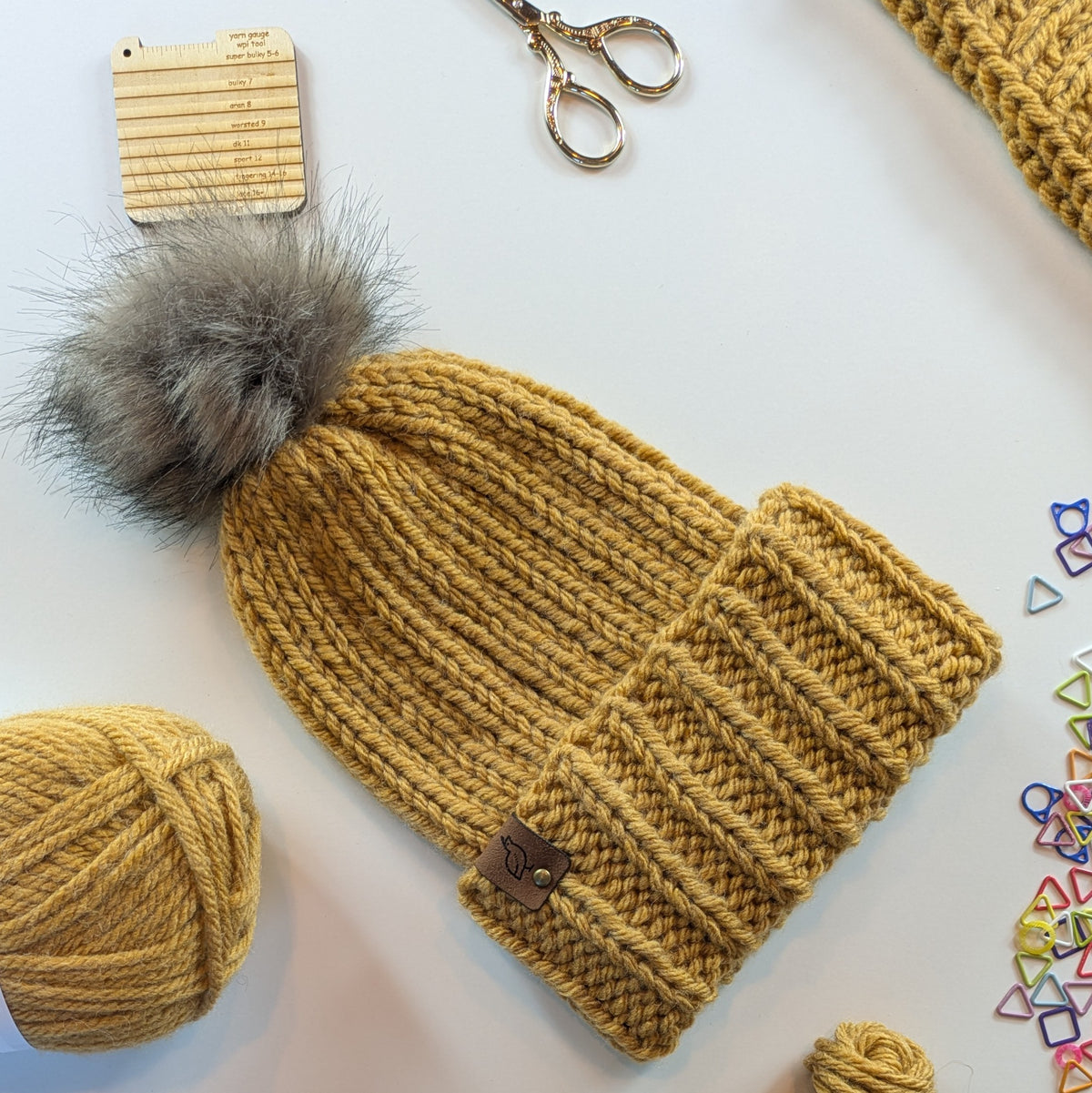 A mustard yellow chunky knit beanie with a large gray faux-fur pom-pom, laid flat on a white surface with knitting supplies nearby including yarn, scissors, a wooden gauge, and small colorful triangle stitch markers.