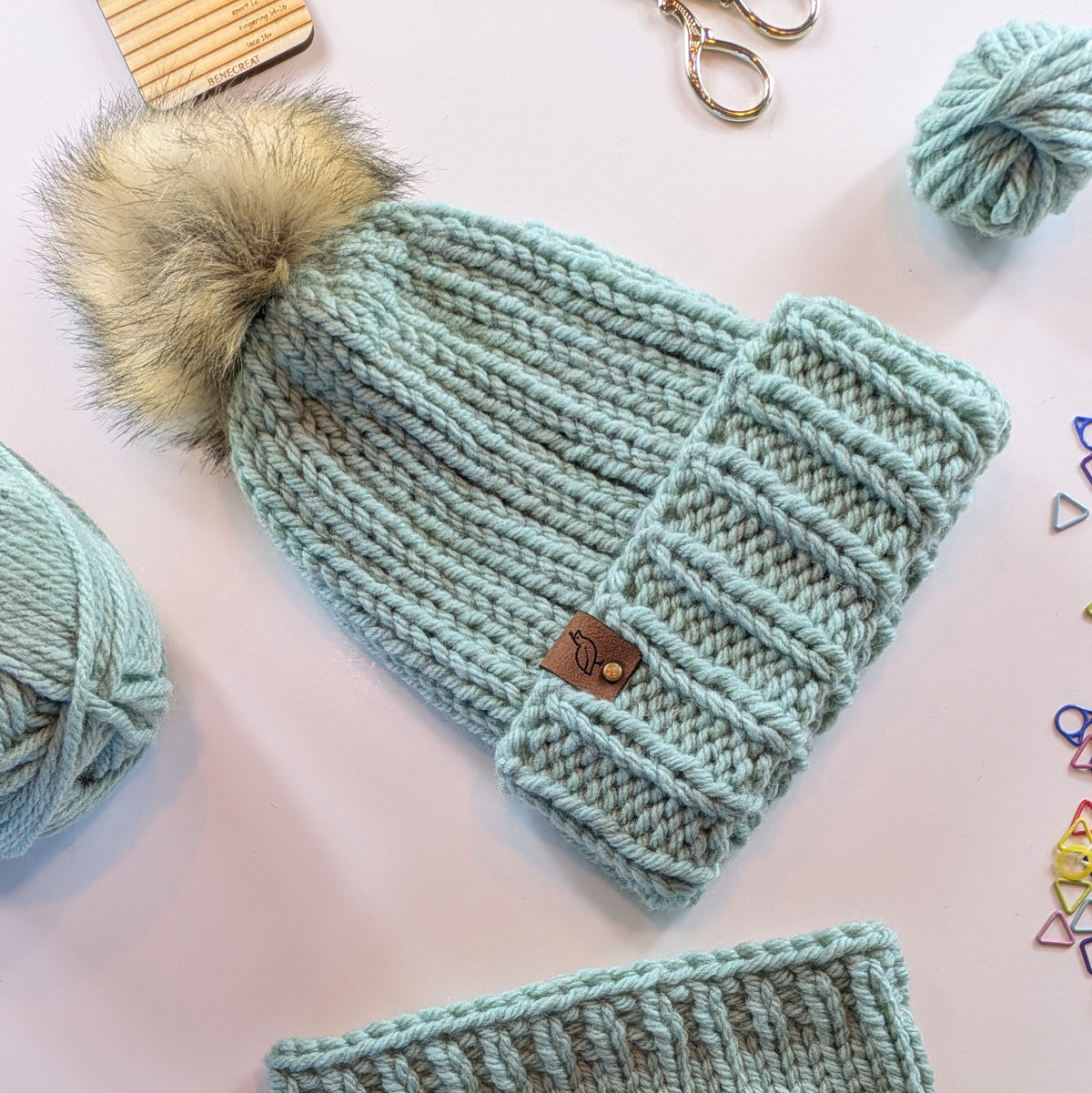 A chunky knit beanie in light mint green with a large beige faux-fur pom-pom, laid flat on a pale pink background with yarn balls, scissors, and colorful stitch markers around it.
