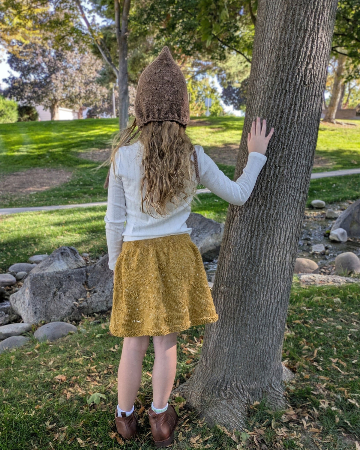 A child seen from behind wearing a brown knit bonnet, a white long-sleeve top, and a mustard yellow skirt, standing outdoors next to a tree in a park.
