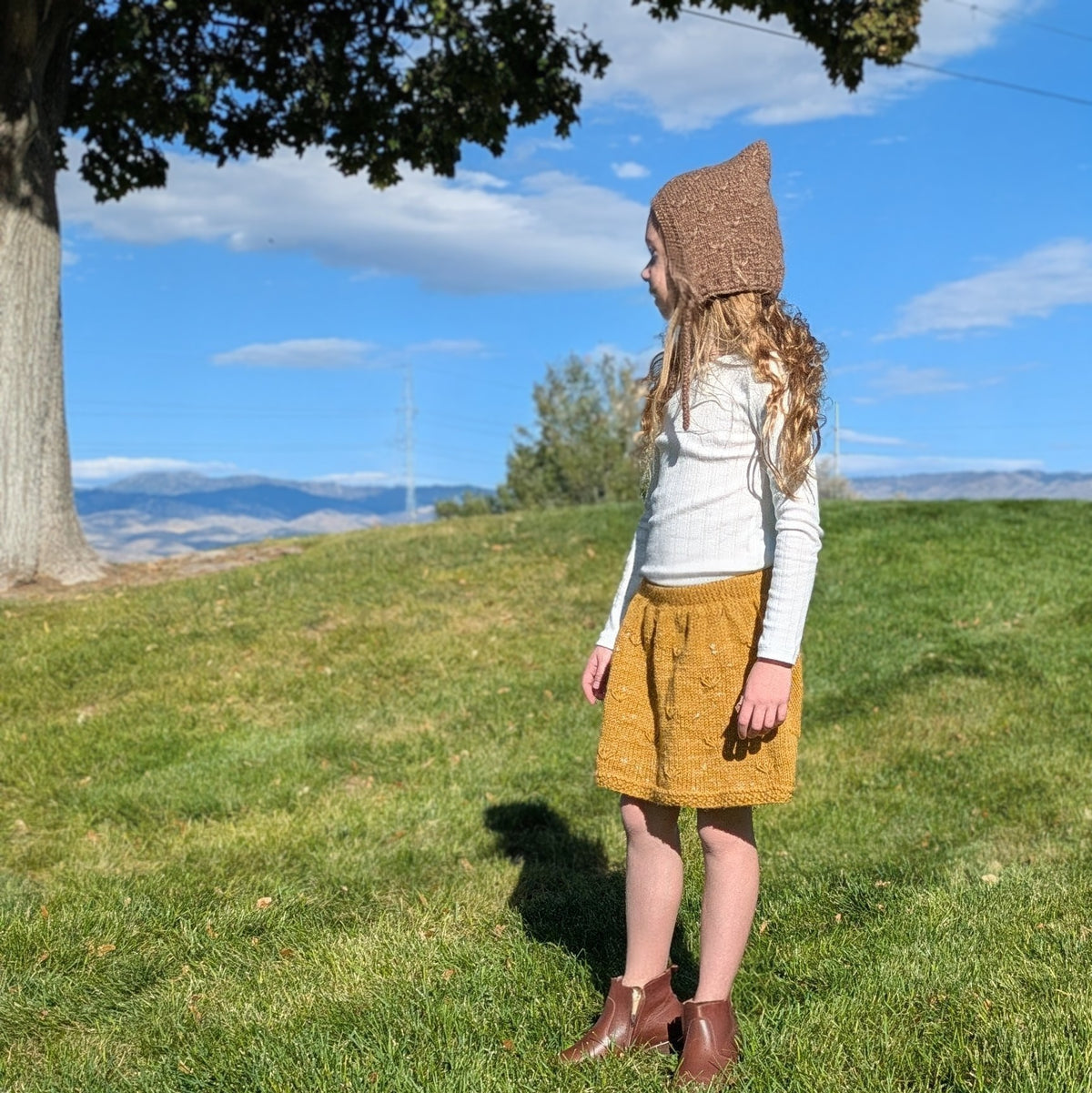 A young girl stands on a grassy hill outdoors wearing a brown knit bonnet, a white long-sleeve top, a mustard yellow hand knit skirt, and brown boots, with a tree, blue sky, and distant hills in the background.
