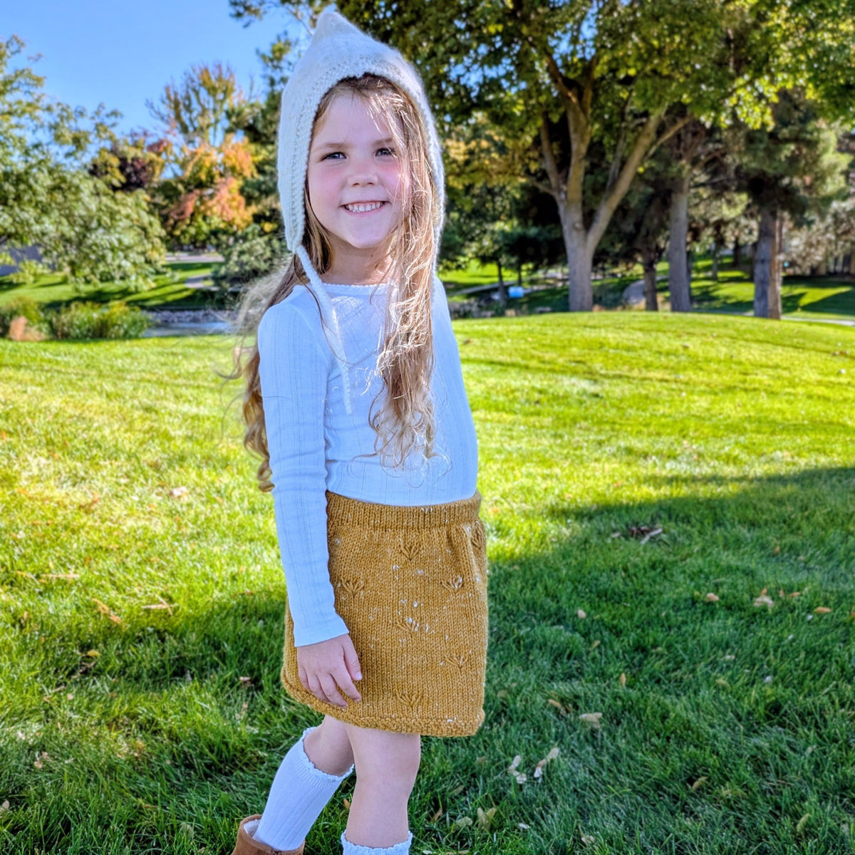 A young girl stands on a grassy park lawn wearing a white long-sleeve top, a mustard yellow knit skirt, white socks, and brown boots, with a white bonnet on.