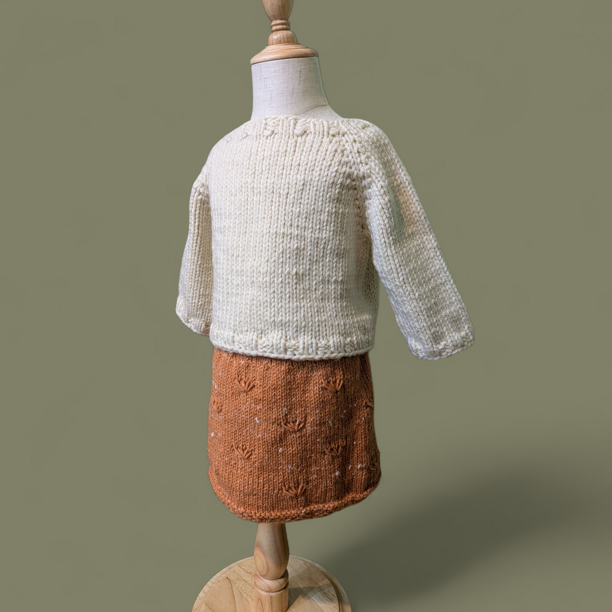 A knitted children's sweater and skirt set displayed on a wooden dress form against a solid sage green background. The sweater is off-white with chunky ribbing at the collar and cuffs, and the skirt is a muted pumpkin orange with a textured dandelion seed pattern.