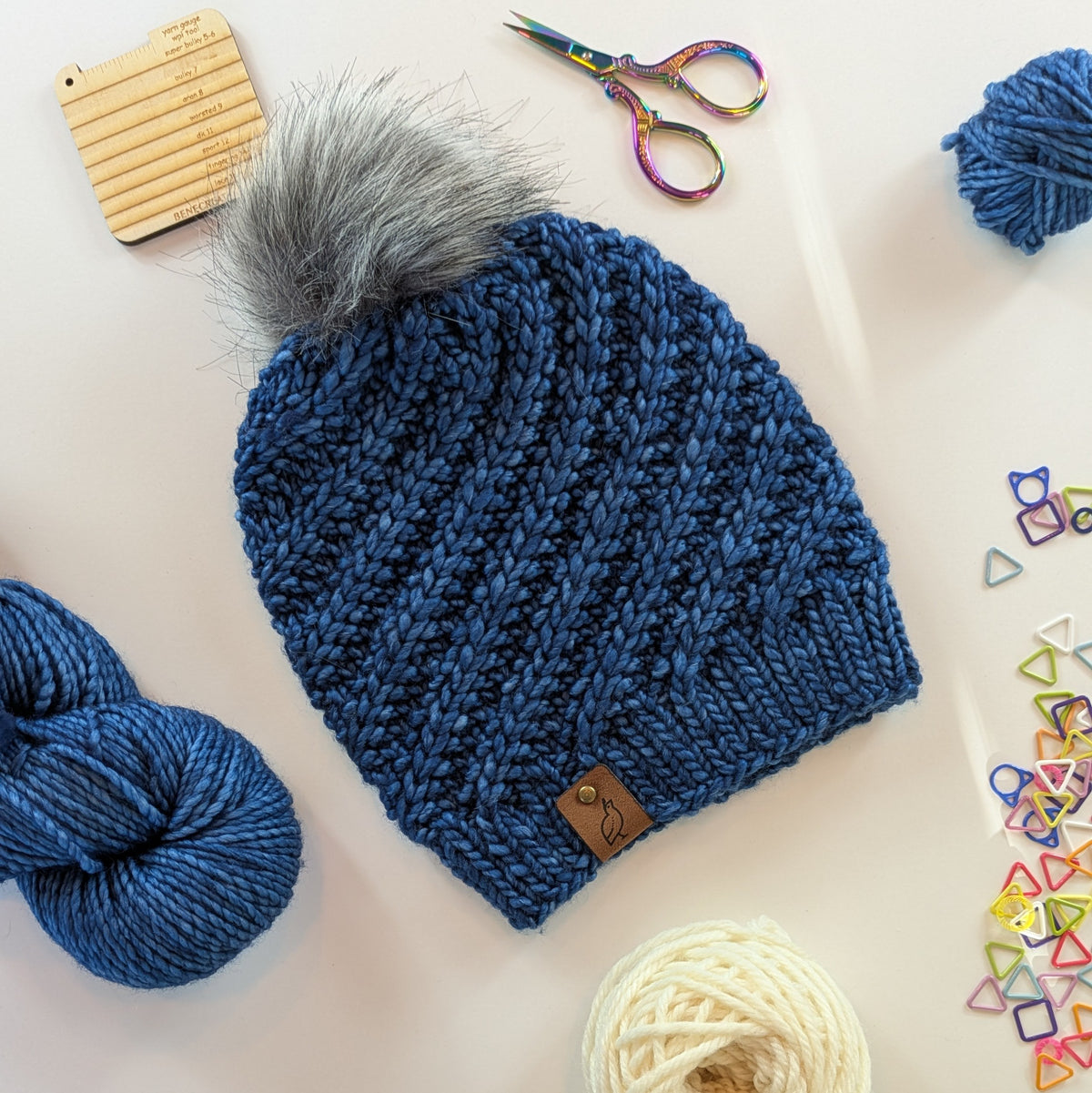A blue chunky knit beanie with a gray faux-fur pom-pom, styled in a flat lay with yarn balls, colorful stitch markers, rainbow scissors, and a wooden stitch sampler on a white background.