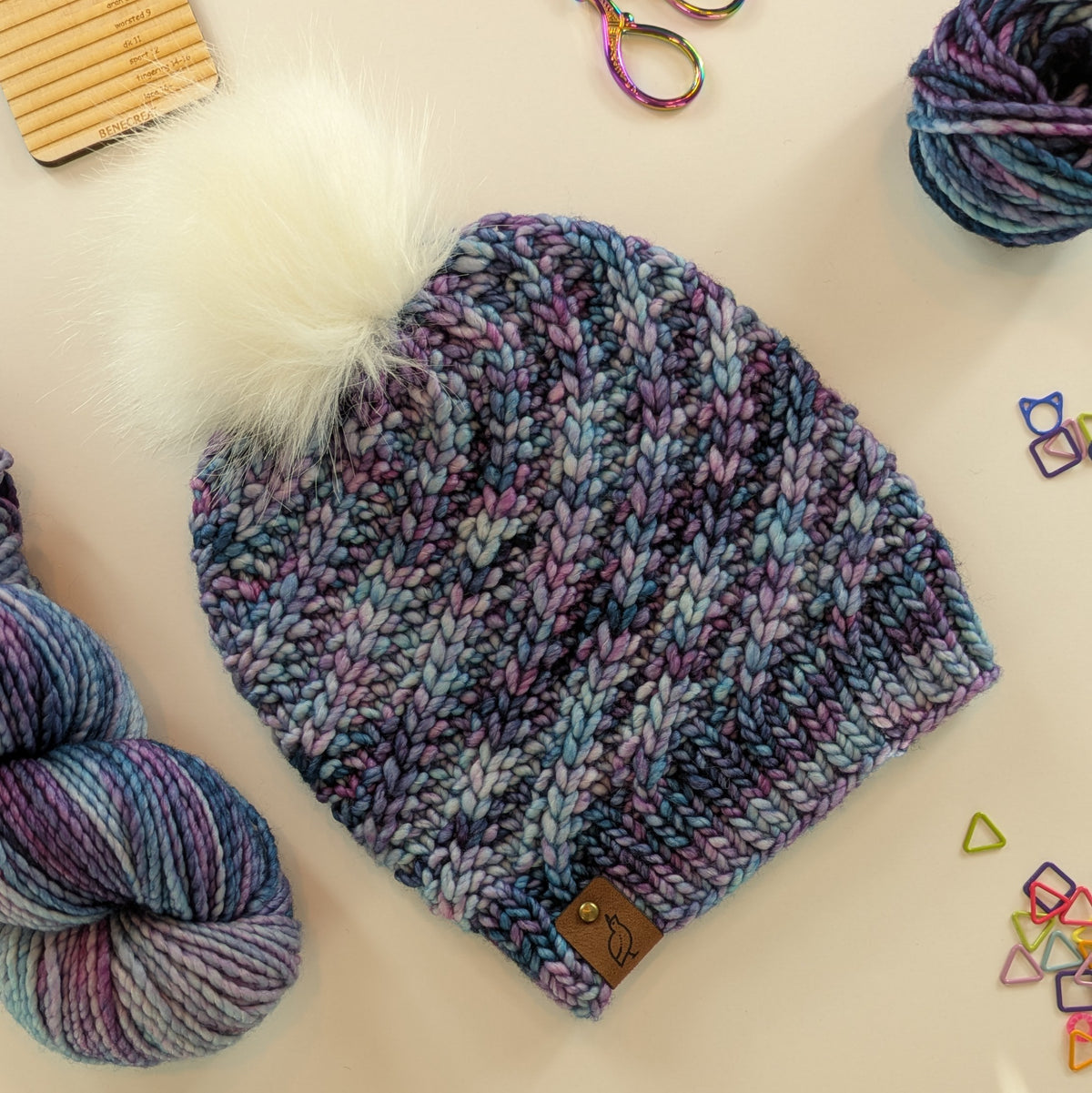 A chunky knit beanie hat in a variegated blend of blue, purple, and gray yarn, topped with a fluffy white pom-pom. The hat is styled in a flat-lay arrangement with skeins of matching variegated yarn, rainbow scissors, and small geometric stitch markers on a light background.