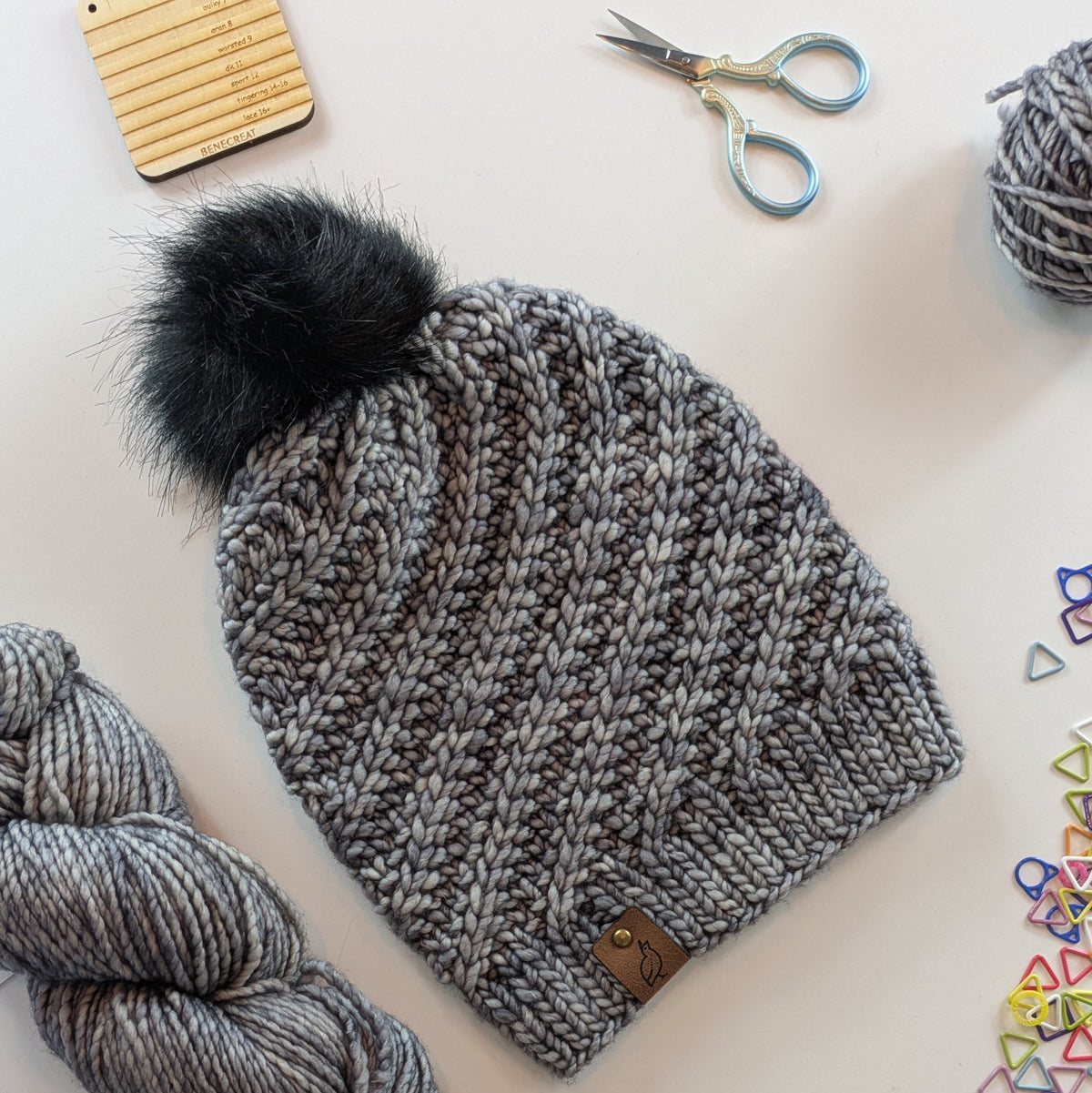 A gray tonal chunky knit beanie with a large black faux fur pom-pom, styled in a flat-lay with knitting supplies including yarn, scissors, and colorful stitch markers on a light background.