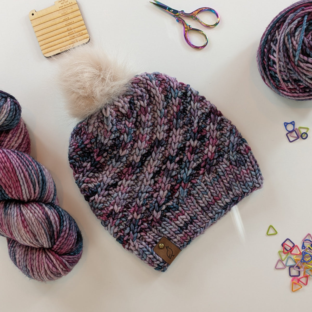 A multicolor variegated knit beanie with a cream faux fur pom-pom and a small brown leather logo tab, styled in a flat-lay with yarn, scissors, and knitting accessories on a white background.