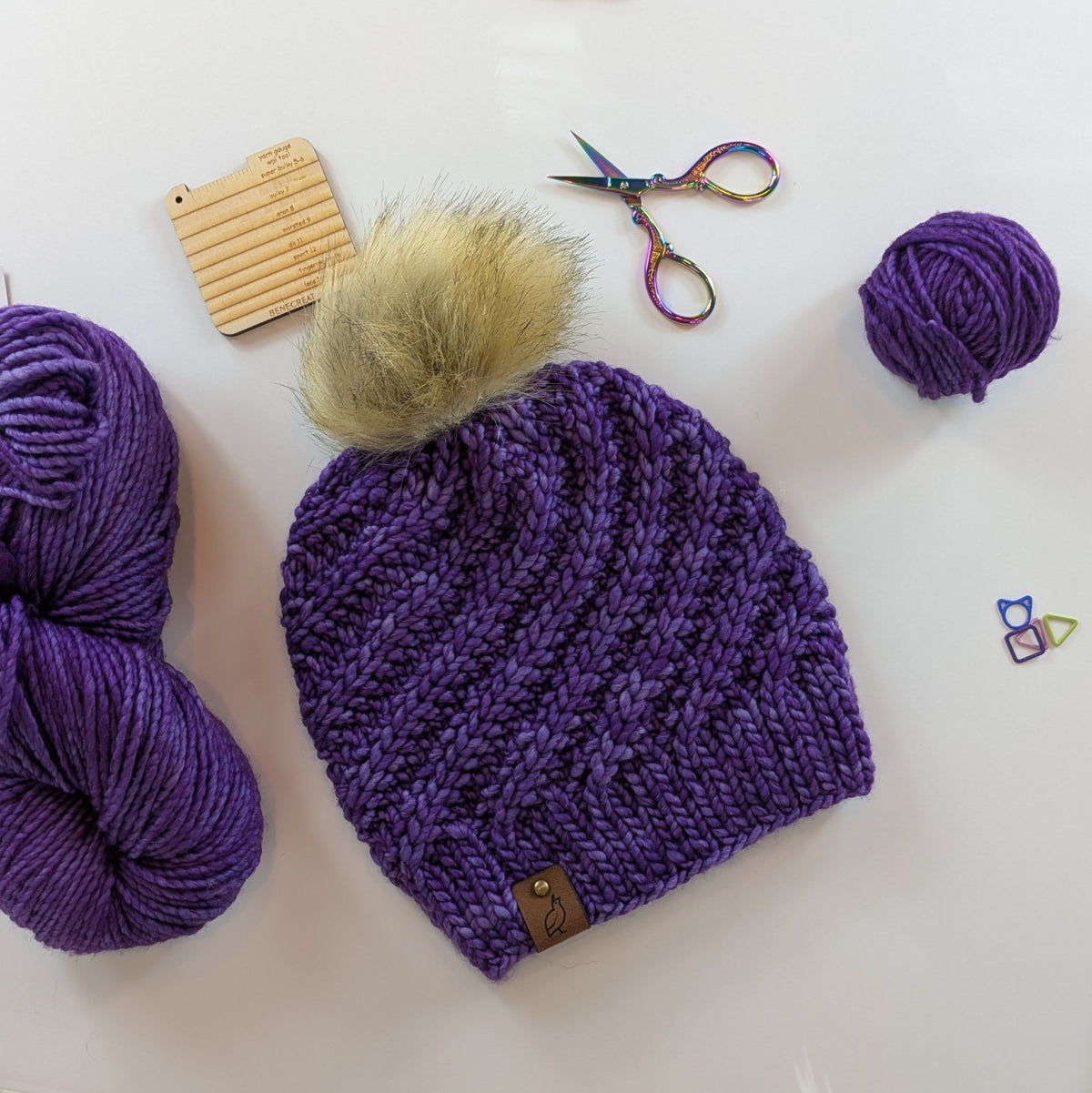 A purple chunky knit beanie with a fluffy beige pom-pom, laid flat on a white surface with knitting supplies including purple yarn balls, a wooden stitch marker, rainbow scissors, and a small colorful triangle stitch marker.