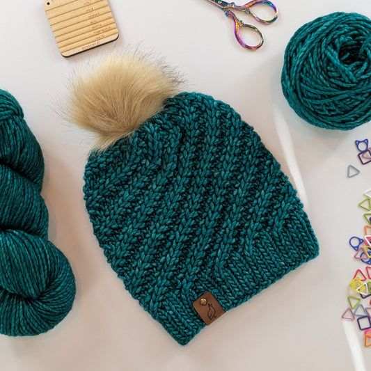 A teal knitted beanie with a large cream faux-fur pom-pom and a small brown leather patch, arranged on a white surface with yarn balls, scissors, and small colorful accessories around it.
