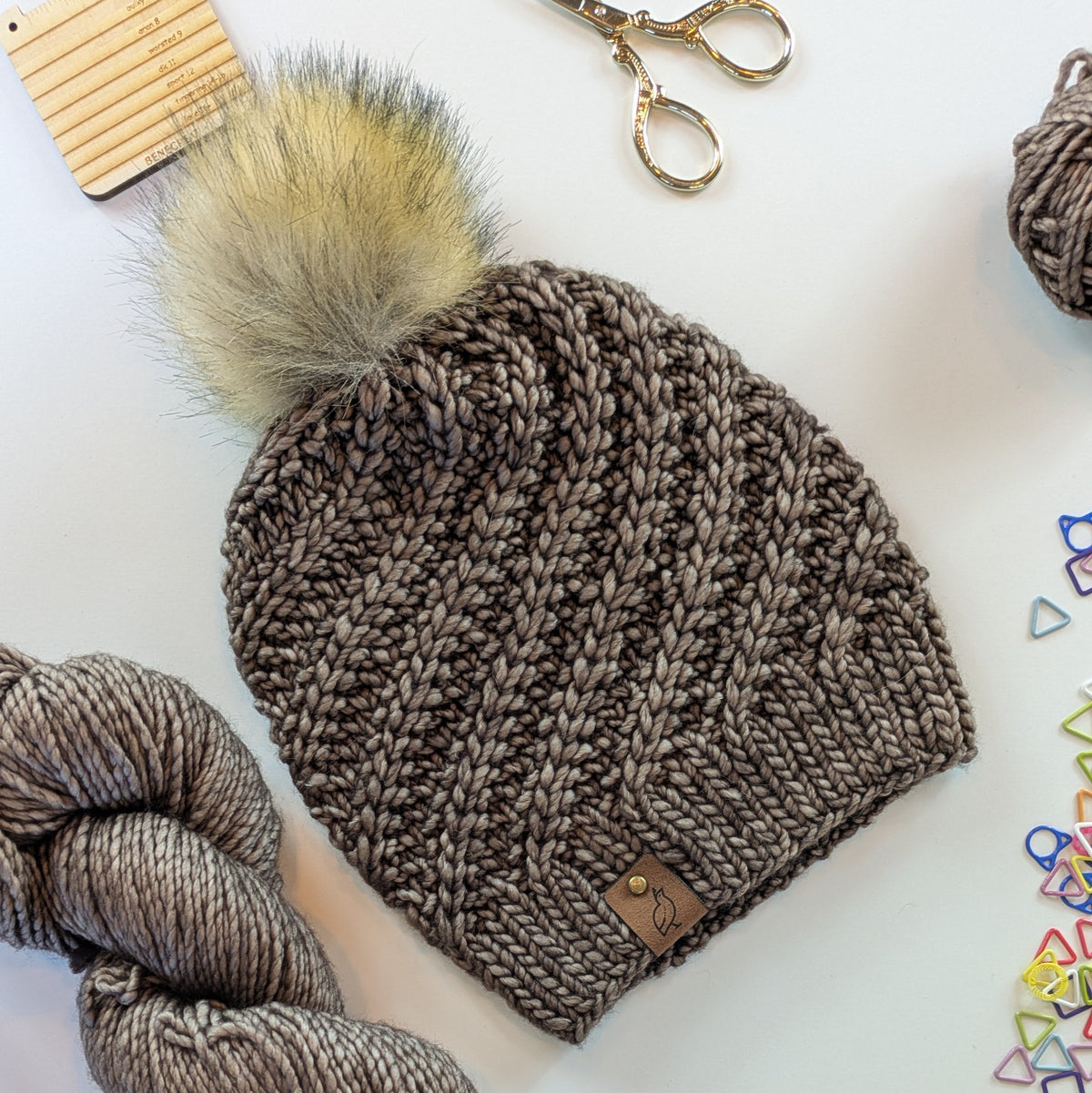 A chunky knit beanie hat in taupe-brown with a large fluffy pom-pom in beige and brown, laid flat on a white surface with knitting props including yarn, scissors, and small colorful triangle clips.