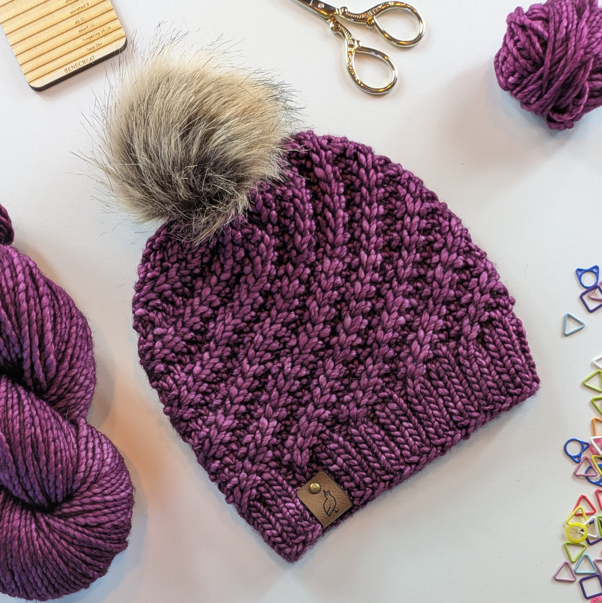 A textured cable-knit beanie in a heathered plum-purple color, featuring a large fluffy faux-fur pom-pom in beige and gray. The beanie is styled in a flat-lay arrangement with purple yarn skeins, gold scissors, and colorful stitch markers on a white background.