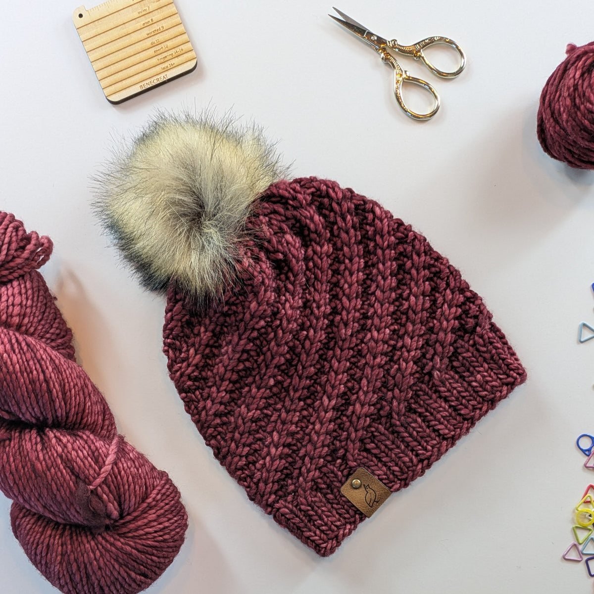 A chunky knit beanie in a deep burgundy/plum color with a large beige-gray faux fur pom-pom, styled in a flat lay with yarn balls, scissors, and a wooden tool on a white background.