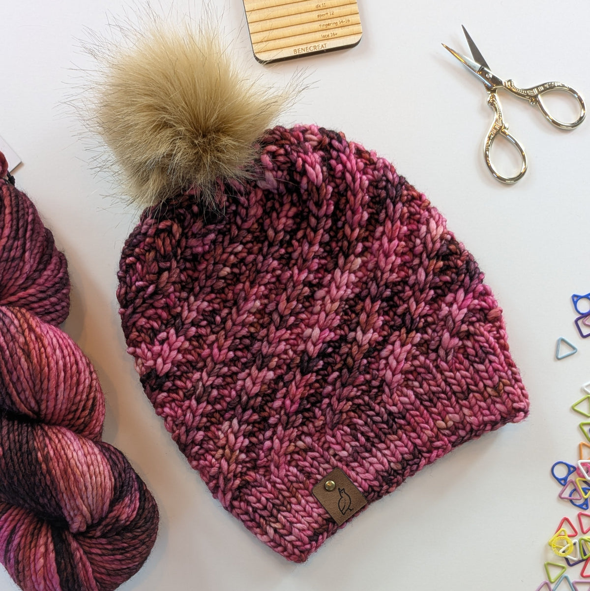 A chunky knit beanie hat in variegated pink, burgundy, and dark tones, topped with a large beige faux-fur pom-pom. The hat is styled in a flat-lay arrangement with a matching variegated skein of yarn, gold scissors, and small colorful triangle clips on a white background.
