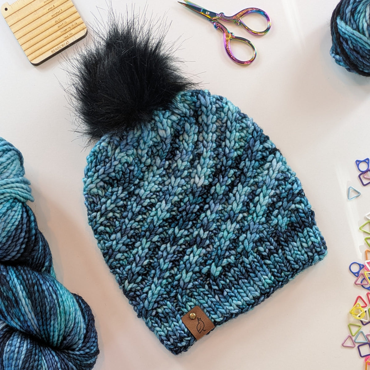 A chunky knit beanie hat in a variegated blue and black yarn, featuring a large black faux fur pom-pom on top and a small brown leather patch with a bronze rivet on the brim. The hat is styled in a flat-lay arrangement with skeins of matching yarn, rainbow scissors, and colorful stitch markers on a white background.
