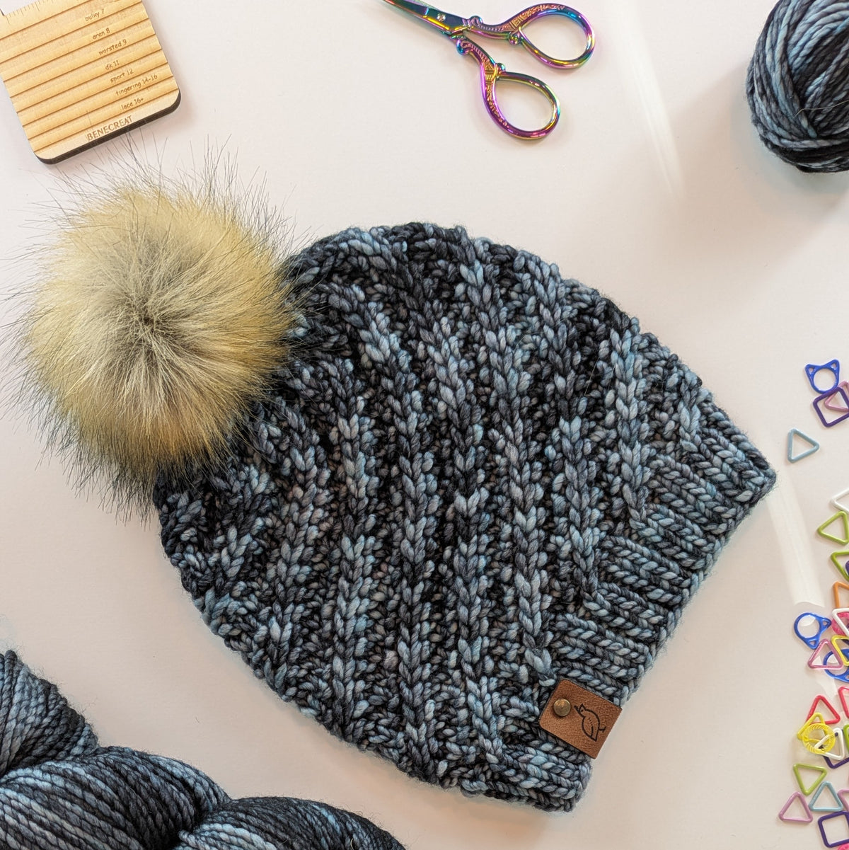 A chunky knit beanie in heathered blue-gray yarn with a large fluffy cream pom-pom, laid flat on a light surface with knitting props around it.
