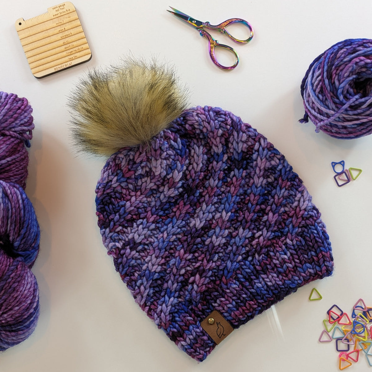 A chunky knit beanie hat in shades of purple, plum, and blue with a large tan faux-fur pom-pom, laid flat on a white surface with yarn balls, scissors, and small colorful stitch markers around it.