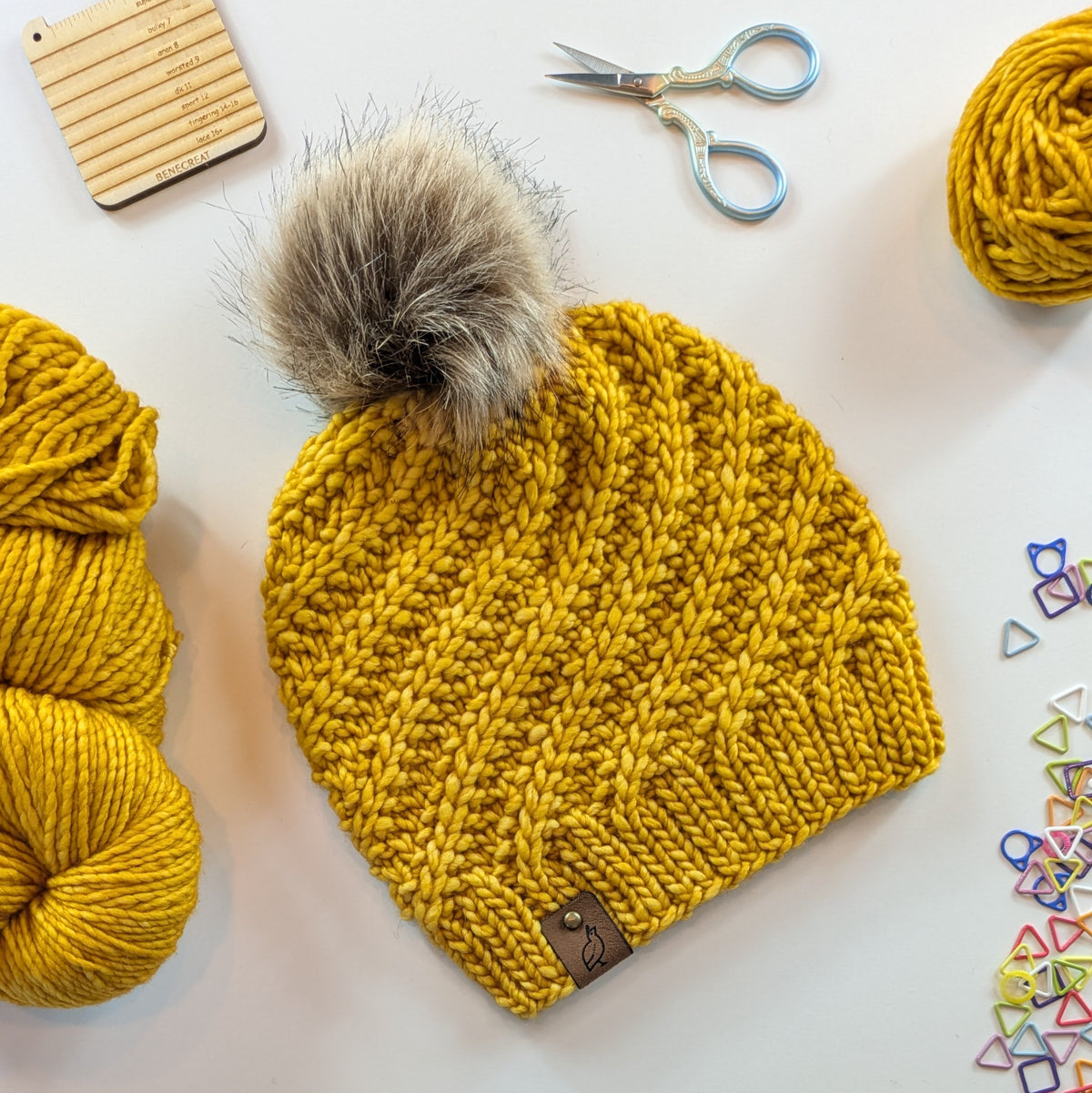 A mustard yellow chunky knit beanie with a large gray-brown faux fur pom-pom, styled in a flat lay with yarn balls, scissors, a wooden ruler, and small colorful geometric clips on a white background.