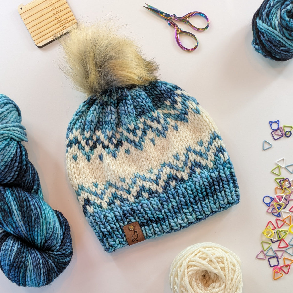 A knitted pom-pom beanie laid flat on a light background, surrounded by yarn balls, scissors, a wooden comb, and small colorful stitch markers. The beanie has a cream and blue fair-isle style pattern with a tan faux-fur pom-pom and a small brown logo tab near the brim.